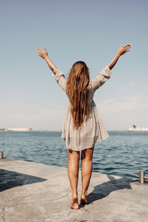 woman in a dress stands on a dock by the water, her arms raised in the air. Concept of freedom and joy, as the woman is celebrating or expressing her happiness.の写真素材