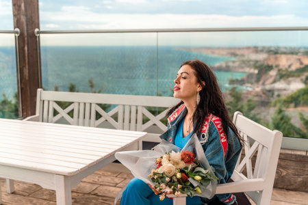A woman sits at a table with a bouquet of flowers in front of her. She is wearing a blue jacket and she is enjoying the view of the ocean.の写真素材