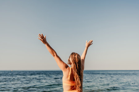A woman with long hair is standing in the sun, with her arms raised in the air. Concept of freedom and joy, as the woman is celebrating or expressing happiness. The bright blue sky.の写真素材
