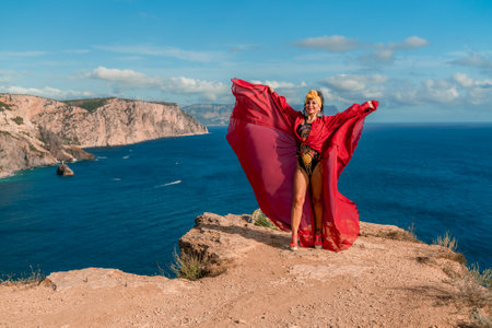 A woman in a red dress is standing on a rocky cliff overlooking the ocean. She is wearing a lei and she is dancing or celebrating. The scene is serene and peaceful, with the ocean as a backdrop.の写真素材