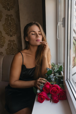 A woman in a black dress is sitting in front of a window with a bouquet of red roses.の写真素材