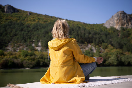 A woman in a yellow jacket is sitting on a blanket by a lakeの写真素材