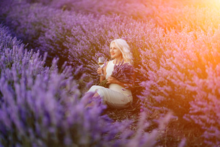 Blonde lavender field holds a glass of white wine in her hands. Happy woman in white dress enjoys lavender field picnic holding a large bouquet of lavender in her hands . Illustrating womans picnic in a lavender field.の写真素材