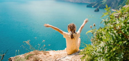 Happy girl perched atop a high rock above the sea, wearing a yellow jumpsuit and braided hair, signifying the concept of summer vacation at the beach.の写真素材