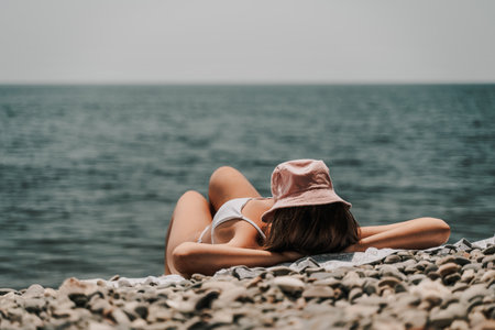 A woman is laying on the beach with a pink hat on her head. She is wearing a white bikini and is laying on a towel. The beach is rocky and the water is calm. Scene is relaxed and peaceful.の写真素材