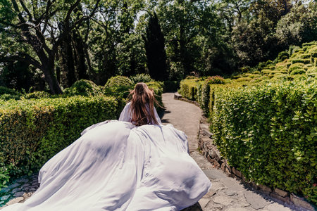 Brunette runs white dress park. A beautiful woman with long brown hair and a long white dress runs along the path along the beautiful bushes in the park, rear viewの写真素材