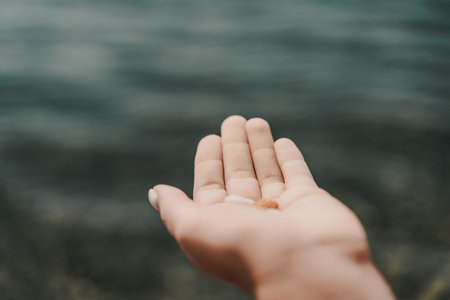 A hand holding a small object in front of a body of water. Concept of calmness and serenity, as the hand is gently holding the object, and the water in the background is calm and still.の写真素材
