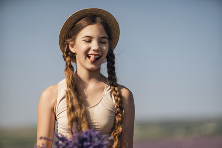 girl is holding a bunch of lavender purple flowers in her hands and wearing a straw hat. She is smiling and she is enjoying herself. The scene is set in a field of lavender, which adds to the peaceful.の写真素材