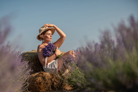woman sitting in a field of lavender and wearing a straw hat. She is smiling and holding a bouquet of flowers. Scene is peaceful and serene, as the woman is surrounded by the beauty of natureの写真素材