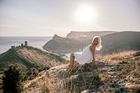 A woman is sitting on a hillside overlooking the ocean. She is wearing a white dress and has blonde hair. The scene is serene and peaceful, with the ocean in the background.の写真素材