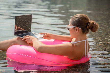 A woman is sitting in a pink inflatable raft on a lake, using a laptop. Concept of relaxation and leisure, as the woman is enjoying her time outdoors while working on her laptop.の写真素材