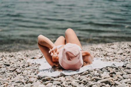 A woman is laying on the beach with a pink hat on her head. She is wearing a white bikini and is laying on a towel. The beach is rocky and the water is calm. Scene is relaxed and peaceful.の写真素材