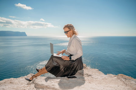 Business woman on nature in white shirt and black skirt. She works with an iPad in the open air with a beautiful view of the sea. The concept of remote work.の写真素材