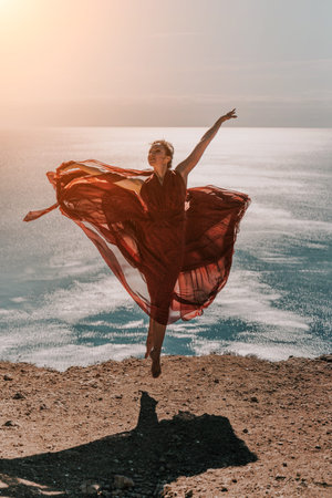 Woman red dress sea. Female dancer in a long red dress posing on a beach with rocks on sunny day. Girl on the nature on blue sky background.の写真素材