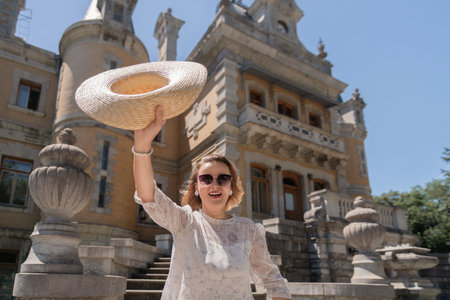A woman is holding a straw hat and smiling in front of a large building