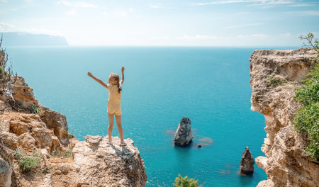 Happy girl stands on a rock high above the sea, wearing a yellow jumpsuit and sporting braided hair, depicting the idea of a summer vacation by the sea.の写真素材