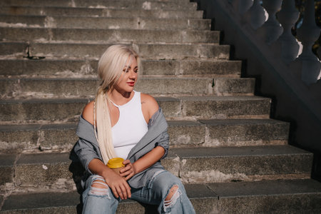 A blonde woman sits on a set of stairs, holding a cup.の写真素材