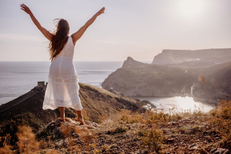 A woman in a white dress stands on a rocky hill overlooking the ocean. She is smiling and she is happy.の写真素材