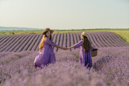 Mom and daughter are running through a lavender field dressed in purple dresses, long hair flowing and wearing hats. The field is full of purple flowers and the sky is clear.の写真素材