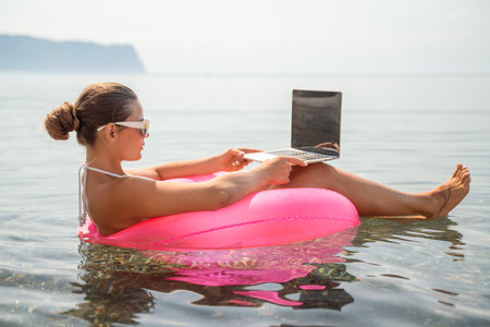 A woman is sitting in a pink inflatable raft on a lake, using a laptop. Concept of relaxation and leisure, as the woman is enjoying her time outdoors while working on her laptop.の写真素材