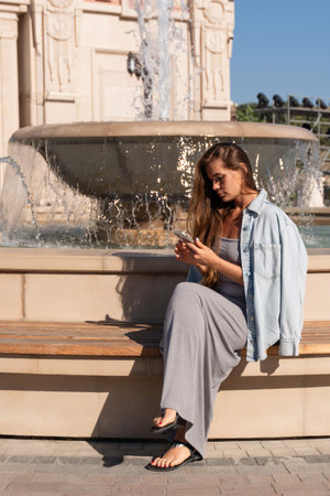 Woman Phone Fountain - Young woman using phone while sitting on a bench next to a fountain in a city.の写真素材