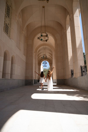 Mosque Courtyard Women Arches - Two women in white dresses walk hand-in-hand through a stone archway in a courtyard.の写真素材