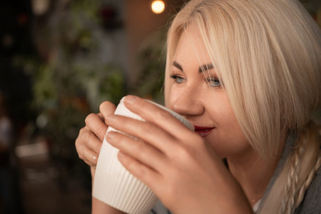 A blonde woman is drinking coffee from a white cup. She is wearing a gray jacket and a necklace.の写真素材