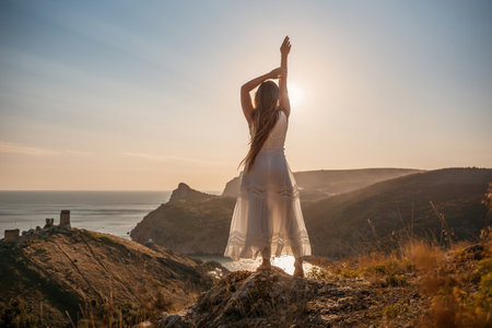 A woman stands on a hill overlooking the ocean. She is wearing a white dress and has her hands on her head. The scene is serene and peaceful, with the woman looking out over the water.の写真素材