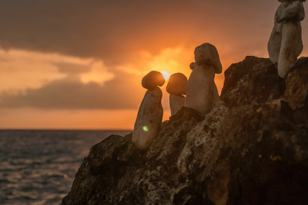 Stone Cairns Sunset Ocean - Silhouettes of balanced rock cairns stand on a cliff against a vibrant sunset over the ocean.の写真素材