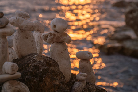 Stone Towers Beach Sunset - Rock sculptures on a beach at sunset. A group of stacked stones, some with faces carved into them, stand on a rocky shore in front of a blurred, golden sunset.の写真素材