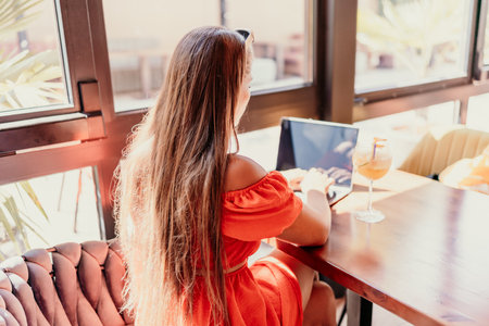 Woman works on laptop in a sunny cafe, glass of juice nearby. The setting is modern, with natural light illuminating the space. The image captures a moment of focused work.の写真素材