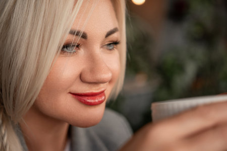 A blonde woman is drinking coffee from a white cup. She is wearing a gray jacket and a necklace.の写真素材