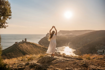 woman stands on a rocky hill overlooking a body of water. She is wearing a white dress and she is in a state of joy or celebration. Concept of freedom and happiness.の写真素材