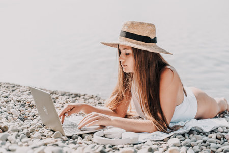 A woman is laying on the beach with a laptop open in front of her. She is wearing a straw hat and a white bikini. Concept of relaxation and leisure.の写真素材
