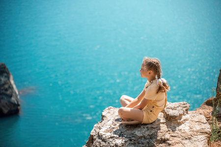 Happy girl perched atop a high rock above the sea, wearing a yellow jumpsuit and braided hair, signifying the concept of summer vacation at the beach.の写真素材