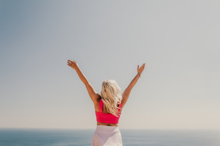 A woman in a pink top and white skirt is standing on a cliff overlooking the ocean. She is smiling and she is happy.の写真素材