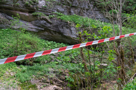 Caution Tape Forest Rockfall Hazard - A red and white caution tape marks a dangerous area in a forest, indicating a rockfall hazard. The tape is stretched across a grassy slope, possibly due to a recent rockfall. The image serves as a warning to hikers and visitors to avoid the area.の写真素材