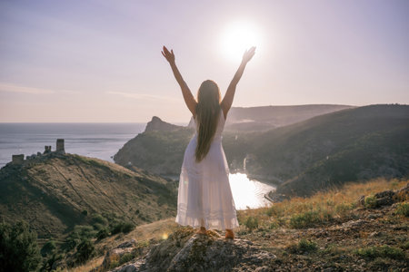 A woman stands on a hill overlooking the ocean. She is wearing a white dress and has her hands on her head. The scene is serene and peaceful, with the woman looking out over the water.の写真素材