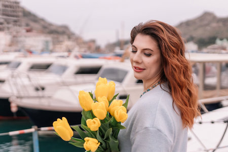 Woman holds yellow tulips in harbor with boats docked in the background., overcast day, yellow sweater, mountainsの写真素材