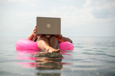 A woman is sitting on a pink inflatable raft in the ocean, using a laptop. Concept of relaxation and...