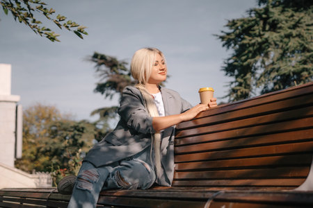 A blonde woman sits on a bench with a cup of coffee in her hand. She is wearing a gray jacket and jeans. The bench is wooden and has a curved back. The scene is peaceful and relaxing.の写真素材