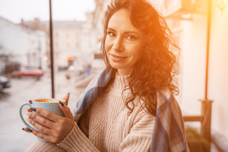 A middle-aged woman in a beige sweater with a blue mug in her hands is in a street cafe on the verandaの写真素材