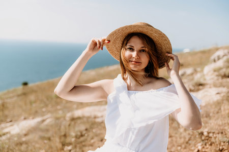 Happy woman in a white dress and hat stands on a rocky cliff above the sea, with the beautiful silhouette of hills in the background.の写真素材