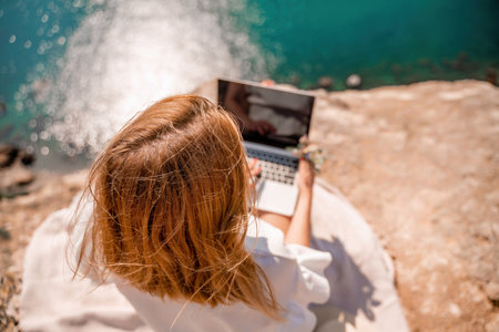 Freelance woman working on a laptop by the sea, typing away on the keyboard while enjoying the beautiful view, highlighting the idea of remote work.の写真素材