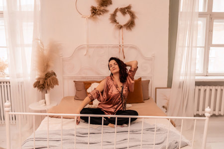 A woman is sitting on a bed with a white frame. She is wearing a pink shirt and is looking at the camera. The room is decorated with white curtains and a white bedspread.の写真素材