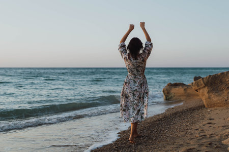 Woman Beach Sunset - A woman wearing a floral dress raises her arms in the air while standing on a sandy beach at sunset.の写真素材