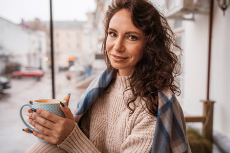 A middle-aged woman in a beige sweater with a blue mug in her hands is in a street cafe on the verandaの写真素材