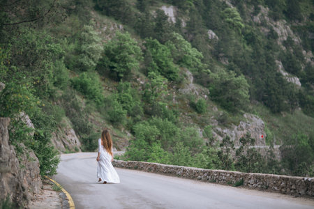 A woman is walking down a road with a beautiful view of the mountains in the background. The scene is serene and peaceful, with the womans dress adding a touch of elegance to the landscape.の写真素材