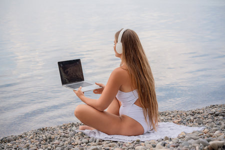 A woman is sitting on the beach with a laptop open in front of her. She is wearing a white bikini and has long hair. Concept of relaxation and leisure.の写真素材