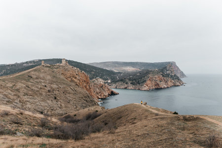 Woman stands on cliff overlooking ocean, cloudy day provides soft lighting. She is wearing a yellow...
